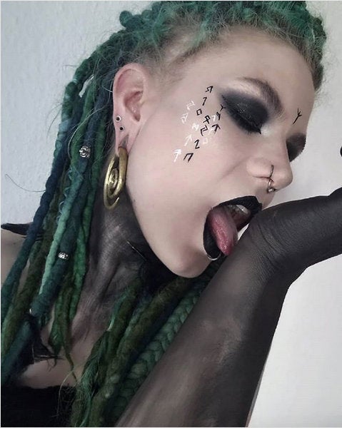 Person with green dreadlocks and dark eye makeup has black paint on their arm and upper body, with multiple piercings, including Spiral Minimalist Ear Hangers in Silver from Kaya Movement at 0 gauge. Sticking their tongue out, touching their hand, Norse runes are painted on their face against a plain, light-colored background.